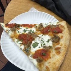 Slice of pizza with ricotta, tomato sauce, sausage, and herbs on a paper plate on a table.