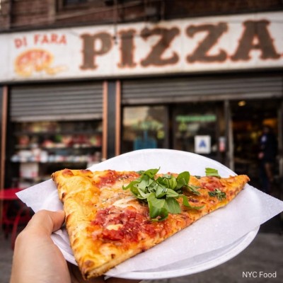 Hand holding a slice of pizza with greens, in front of a pizza restaurant.