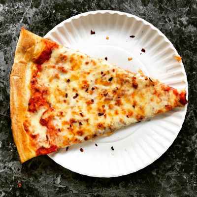 Cheese pizza slice with red pepper flakes on a paper plate.