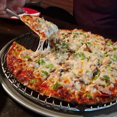 Hand holding slice of veggie pizza with cheese pull on metal tray.