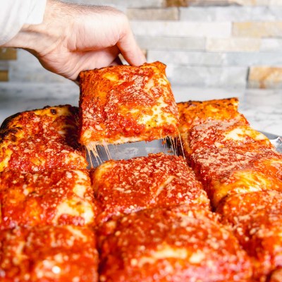 Hand lifting a slice of cheesy, square pizza with tomato sauce from a tray.