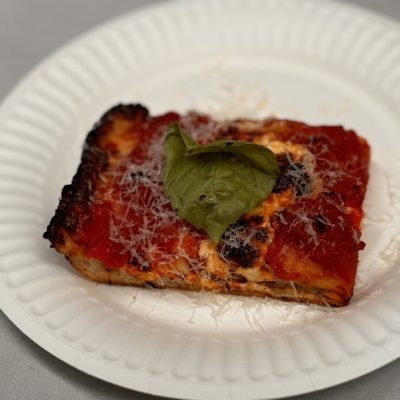 Square slice of pizza on a paper plate, topped with tomato sauce, grated cheese, and basil.
