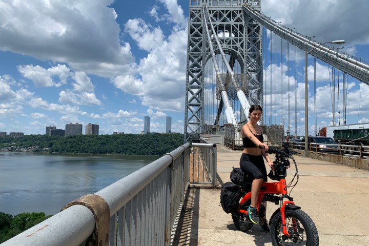 Person on an orange e-bike on a bridge with cloudy sky and river view.