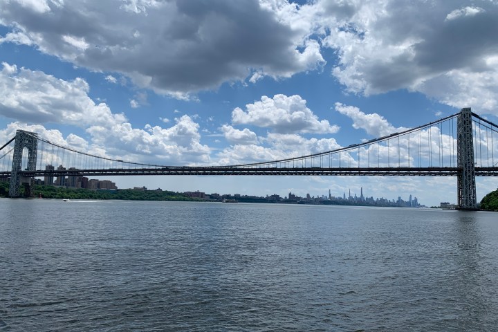 Suspension bridge over a river with city skyline and cloudy sky.