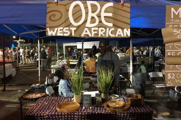 A vendor at a West African food stall under a blue tent at an outdoor event.