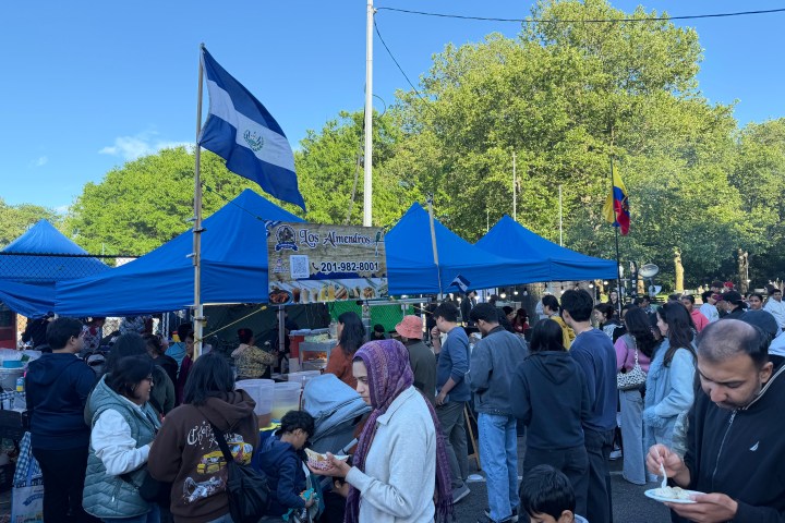 Crowd at outdoor market with blue tents and a flag, people eating and shopping.