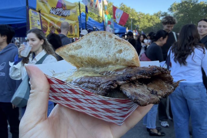 Hand holding a sandwich at an outdoor event with people and booths in the background.