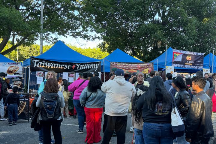 People gathered at outdoor food stalls with blue tents and trees in background.