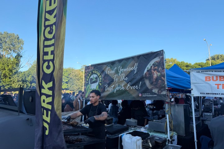 Man grilling at food stall with 'Jerk Chicken' sign under clear sky.