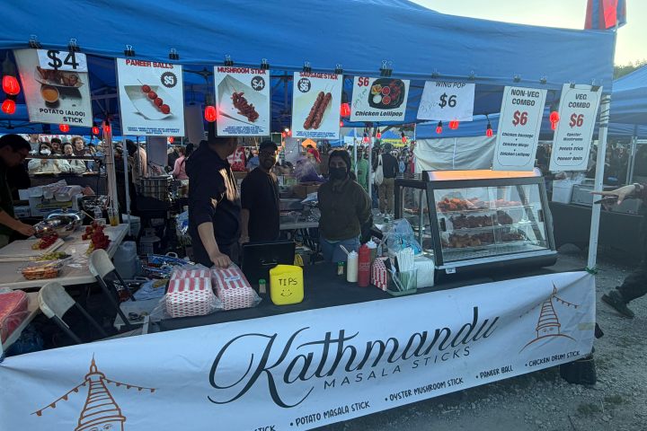 Outdoor food stall with Kathmandu Masala sticks, menu signs, and people behind the counter.