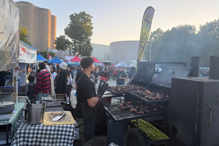 Outdoor event with a man grilling meat at a food stall, smoke rising, and people in the background.